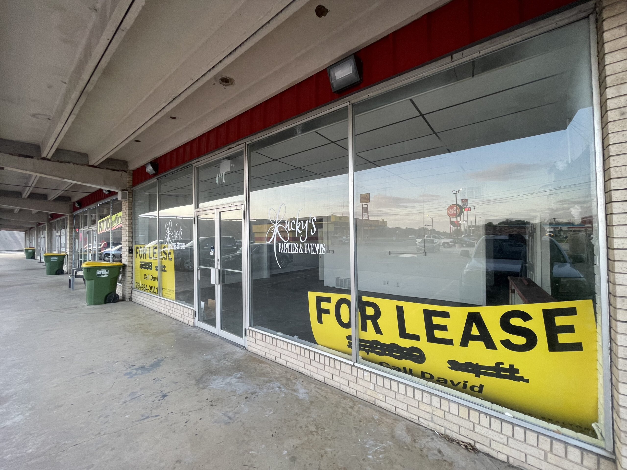 exterior facade under awning of retail space with large windows and doors with brick outline and red accents with yellow for lease sign in window